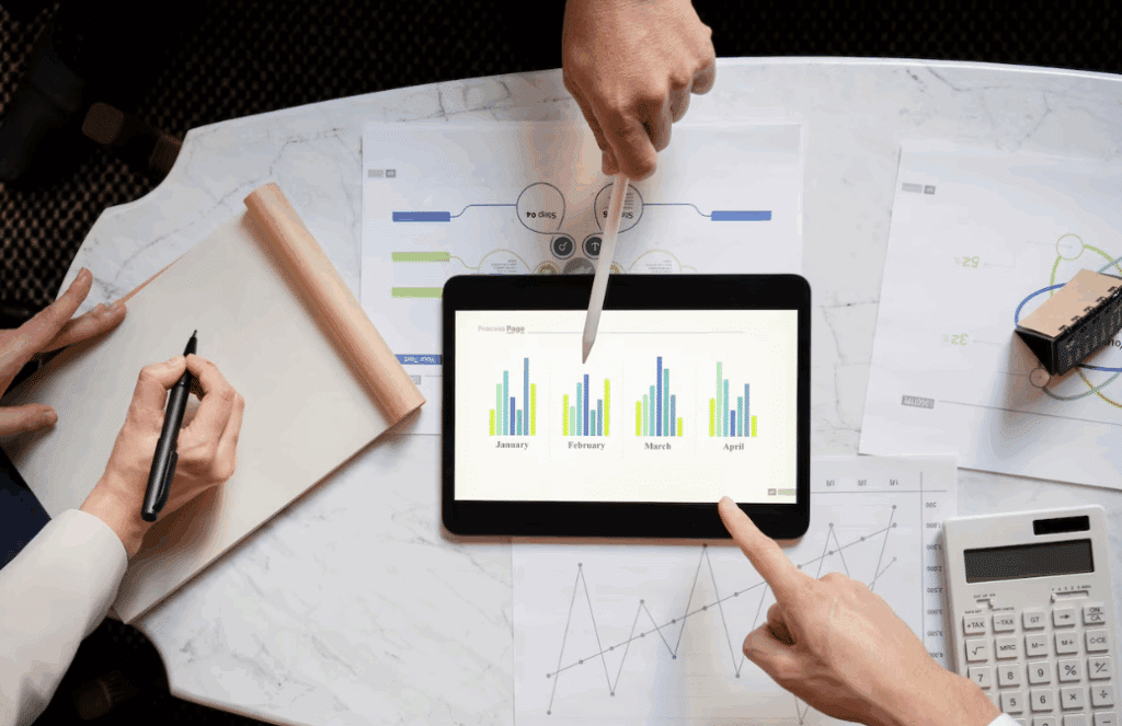 Overhead view of hands analyzing a bar chart on a tablet during a business meeting, with notepads, printed data, and a calculator on a white marble table.