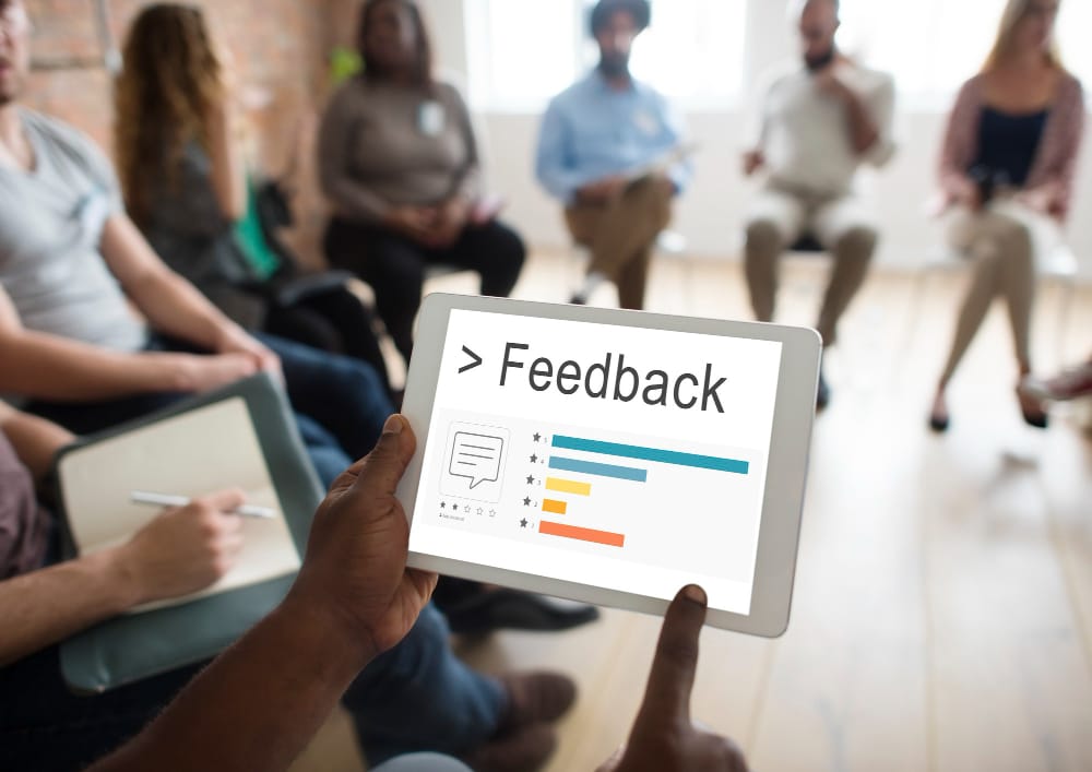 A person holds a tablet displaying "Feedback" and a related graphic, while a blurred group sits in the background, suggesting a feedback session.