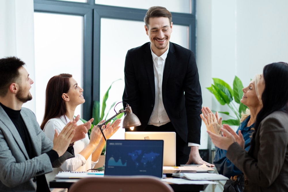 A man in a suit is being applauded by his diverse colleagues at a meeting, where a laptop displays a world map.