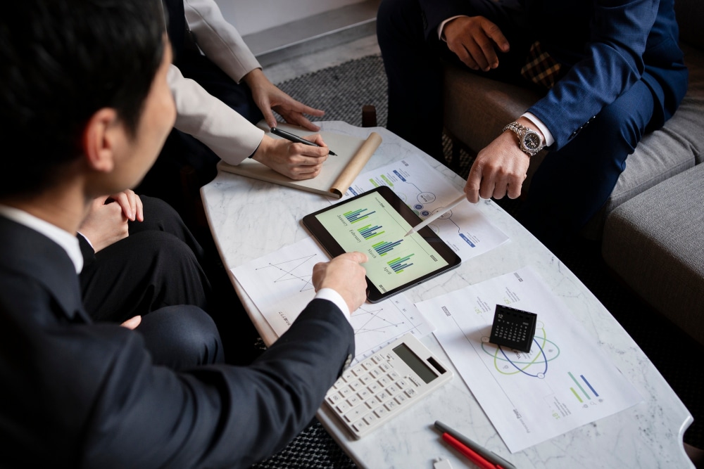 Business team in formal attire sitting around a table analyzing bar charts on a tablet, with printed data sheets, a calculator, and a notepad in view.