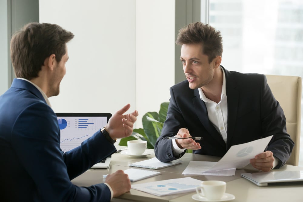 Two businessmen in a discussion at an office desk, one holding a pen and paper while speaking, with a laptop displaying graphs and a coffee cup visible on the table.