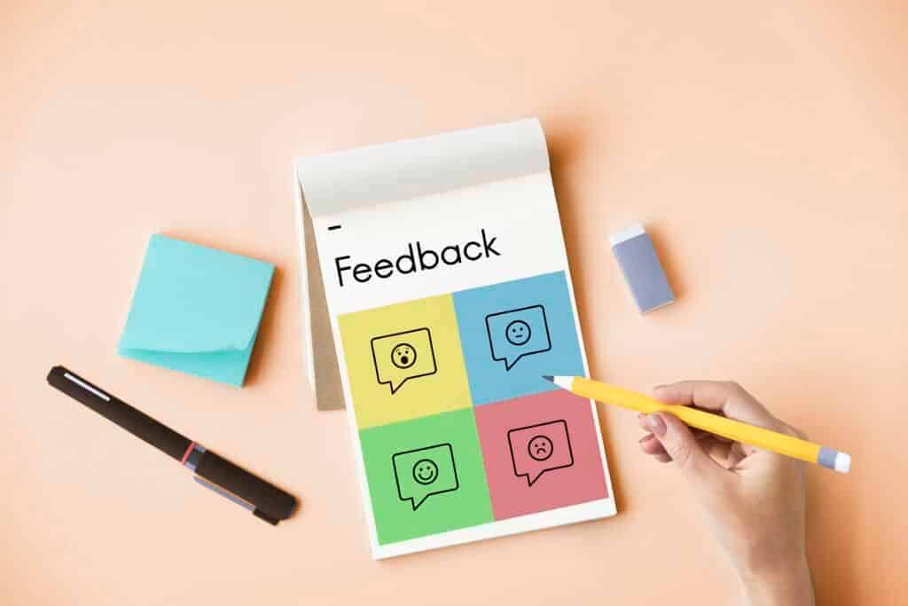**Alt Text:**
A person holding a pencil next to a feedback notebook with colorful emoji icons representing different emotions, symbolizing employee feedback and performance evaluation.
