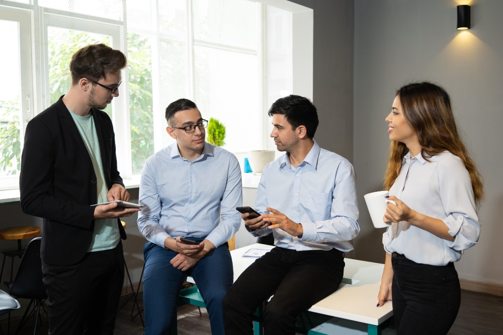 Four coworkers having a casual discussion in a modern office setting, holding a tablet, phones, and a coffee mug.