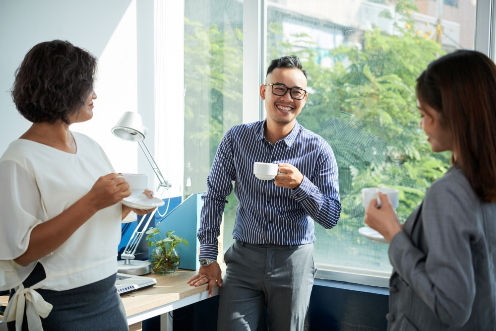 Three colleagues standing by a window, smiling and having a casual conversation while holding coffee cups in a bright office setting.
