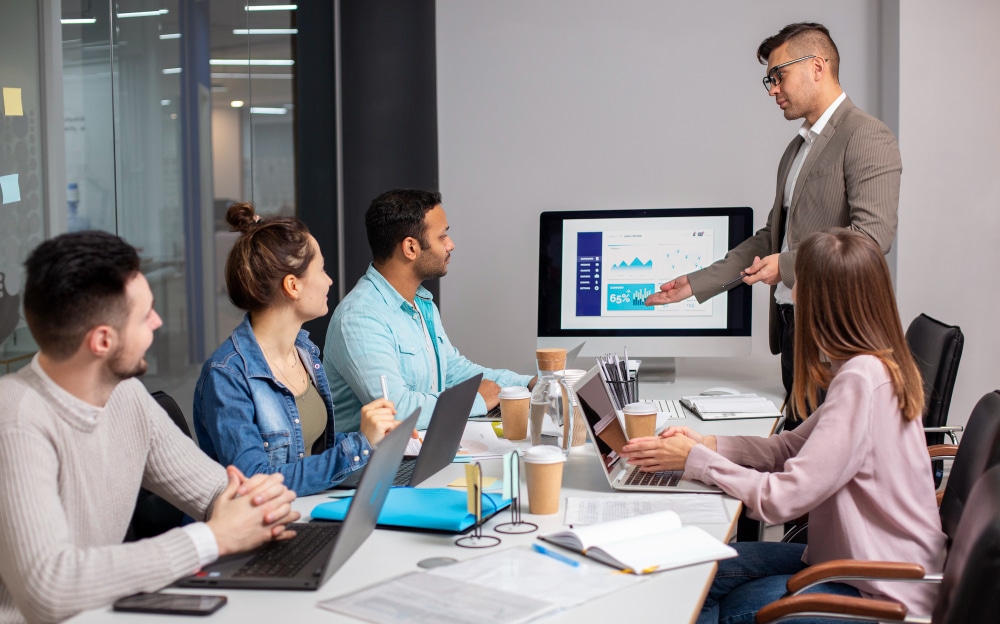 A business team in a modern conference room attending a presentation. A man in a suit is standing and pointing at a screen displaying charts and analytics, while colleagues seated around the table engage with laptops, notebooks, and coffee cups.