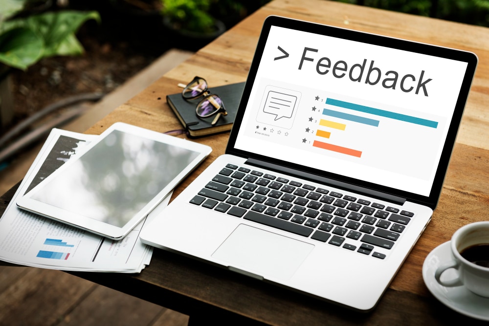 Laptop screen displaying a feedback dashboard with bar charts and a comment icon. The word "Feedback" is prominently shown. Nearby, a tablet, documents with graphs, eyeglasses, and a coffee cup sit on a wooden table, suggesting a work or evaluation setting.