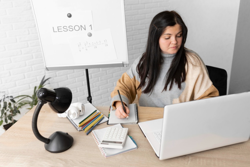 Woman studying at desk with laptop, notebooks, and stationery, in front of a whiteboard with math.
