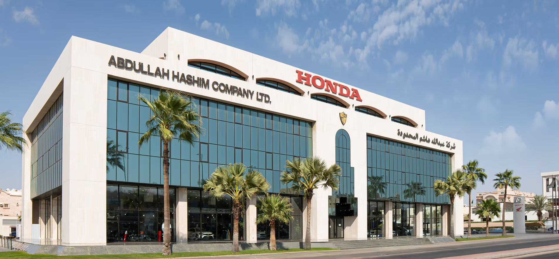 Wide exterior shot of the Abdullah Hashim Company Ltd. headquarters and Honda showroom in Saudi Arabia, featuring a modern multi-story glass facade, palm trees, and clear blue skies.