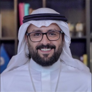 Smiling man wearing glasses and traditional white Gulf attire with a black headband, photographed against a blurred bookshelf background.