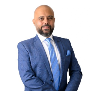Smiling bald man with a beard wearing a blue plaid suit, striped blue tie, and pocket square, photographed against a plain white background.