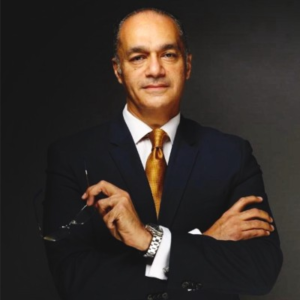 Man in a dark navy suit with a gold tie standing with arms crossed, holding glasses, photographed against a dark gray background.