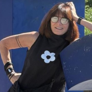 Smiling woman with short auburn hair wearing sunglasses and a black sleeveless top with a white flower design, posing outdoors against a blue backdrop.