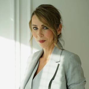 Woman with hair pulled back wearing a light gray blazer, looking at the camera in a bright, softly lit room.