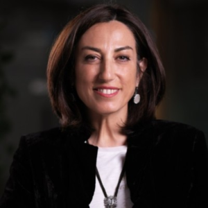 Smiling woman with shoulder-length dark hair wearing a black jacket over a white top and a pendant necklace, photographed against a dark background.