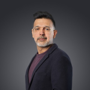 Man with glasses and a short beard wearing a dark navy blazer over a maroon shirt, photographed against a gray gradient background.