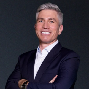 Smiling man with gray hair wearing a dark navy suit over a light blue shirt, standing with arms crossed against a dark background.