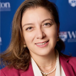 Smiling woman with long brown hair wearing a magenta blazer and pearl necklace, photographed against a blue backdrop with partial logos.
