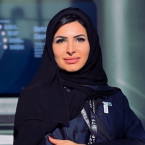 Woman wearing a black hijab and abaya with a lanyard, seated in front of a blue-lit corporate display.