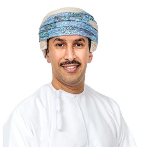 Smiling man wearing a white dishdasha and a patterned Omani mussar turban, photographed against a plain white background.