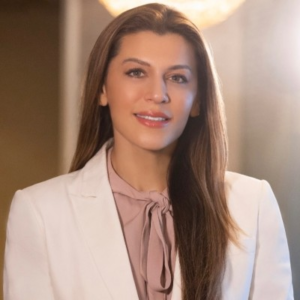 Smiling woman with long dark hair wearing a white blazer over a pink pussy-bow blouse, photographed in a softly lit interior.
