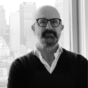 Black-and-white photo of a bald man with a beard and glasses wearing a dark sweater over a white shirt, standing in front of a city skyline.