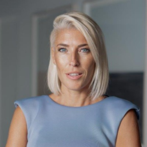 Woman with short blonde hair wearing a light blue top, looking at the camera in a softly lit modern interior.