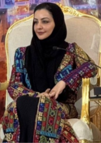 Woman wearing a black headscarf and a colorful embroidered abaya, seated in an ornate gold-framed chair at an event.