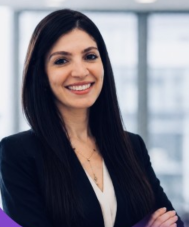 Smiling woman with long dark hair wearing a black blazer over a white top, standing with arms crossed in a bright modern office.