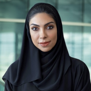 Woman wearing a black hijab and abaya, looking at the camera against a softly blurred green-toned modern background.