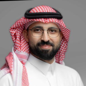 Man with glasses and a beard wearing traditional Gulf attire with a red-and-white checkered shemagh and black agal, against a gray background.