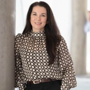 Smiling woman with long dark hair wearing a patterned blouse and black trousers, standing beside a concrete pillar in a bright interior.