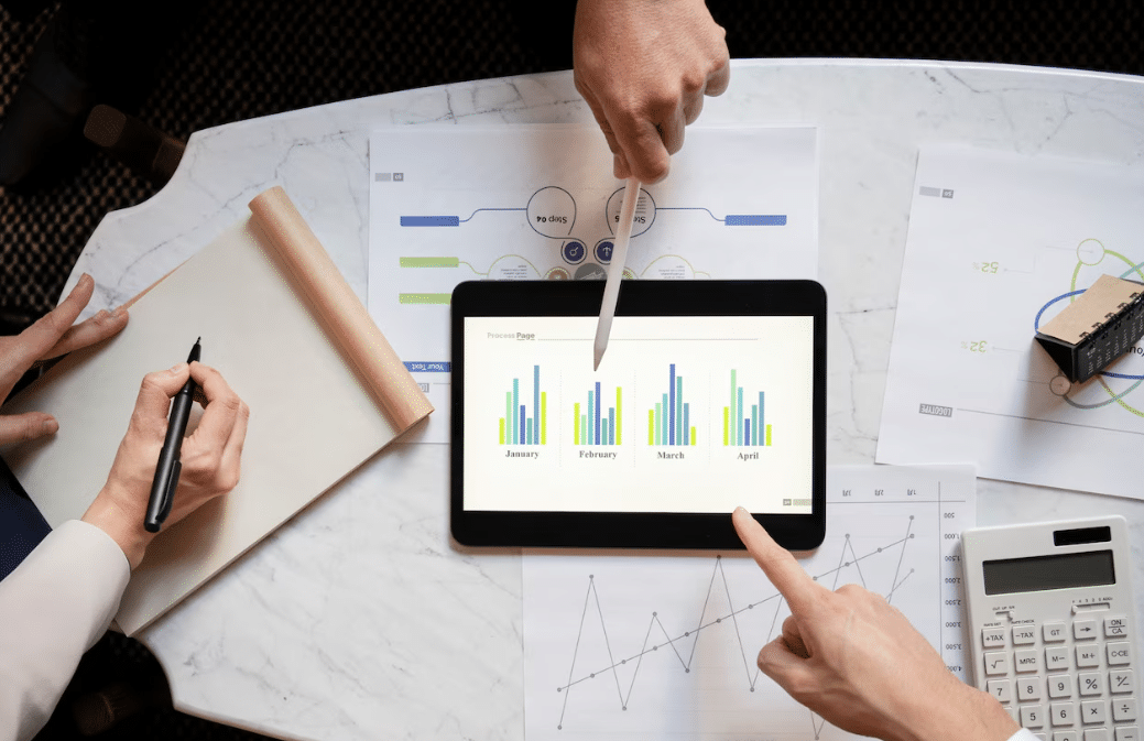 Overhead view of hands analyzing a bar chart on a tablet during a business meeting, with notepads, printed data, and a calculator on a white marble table.
